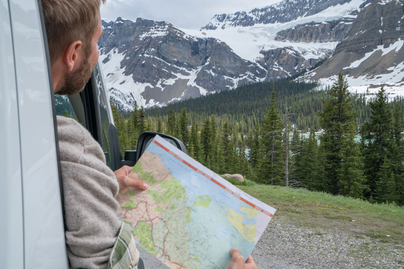 man leaning out side of vehicle with road map in his hands, mountains in the distance