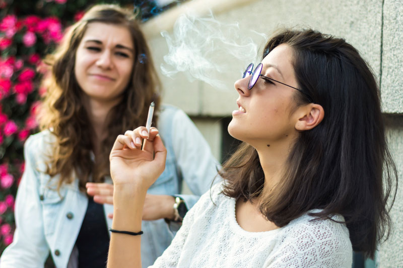 Girl smoking in foreground, with another girl with a look of disgust on her face in the background